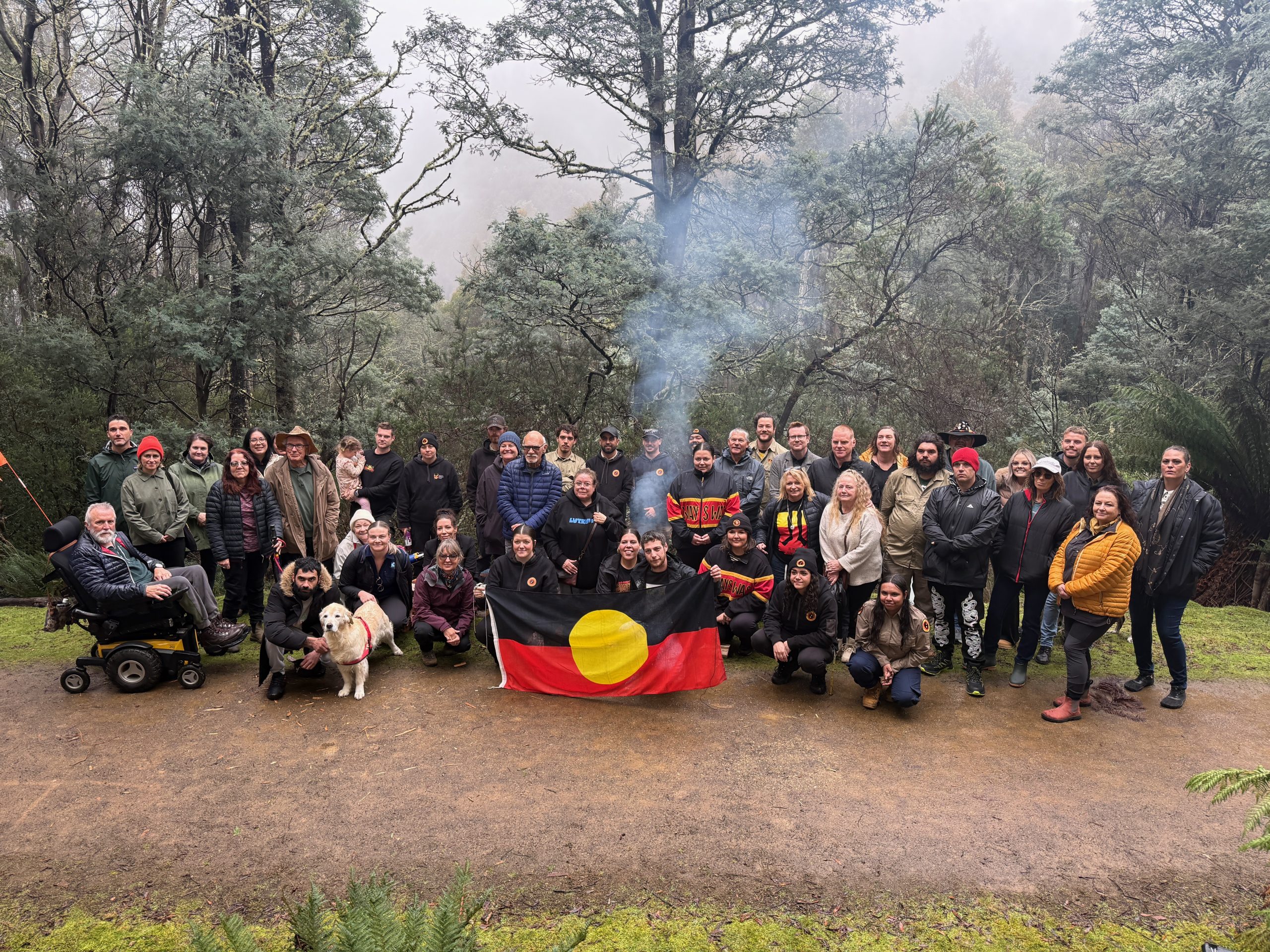 A group of Aboriginal and non-Aboriginal people are photographed behind an Aboriginal Flag. They are standing on a dirt path with a backdrop of trees, mist and smoke from a fire.
